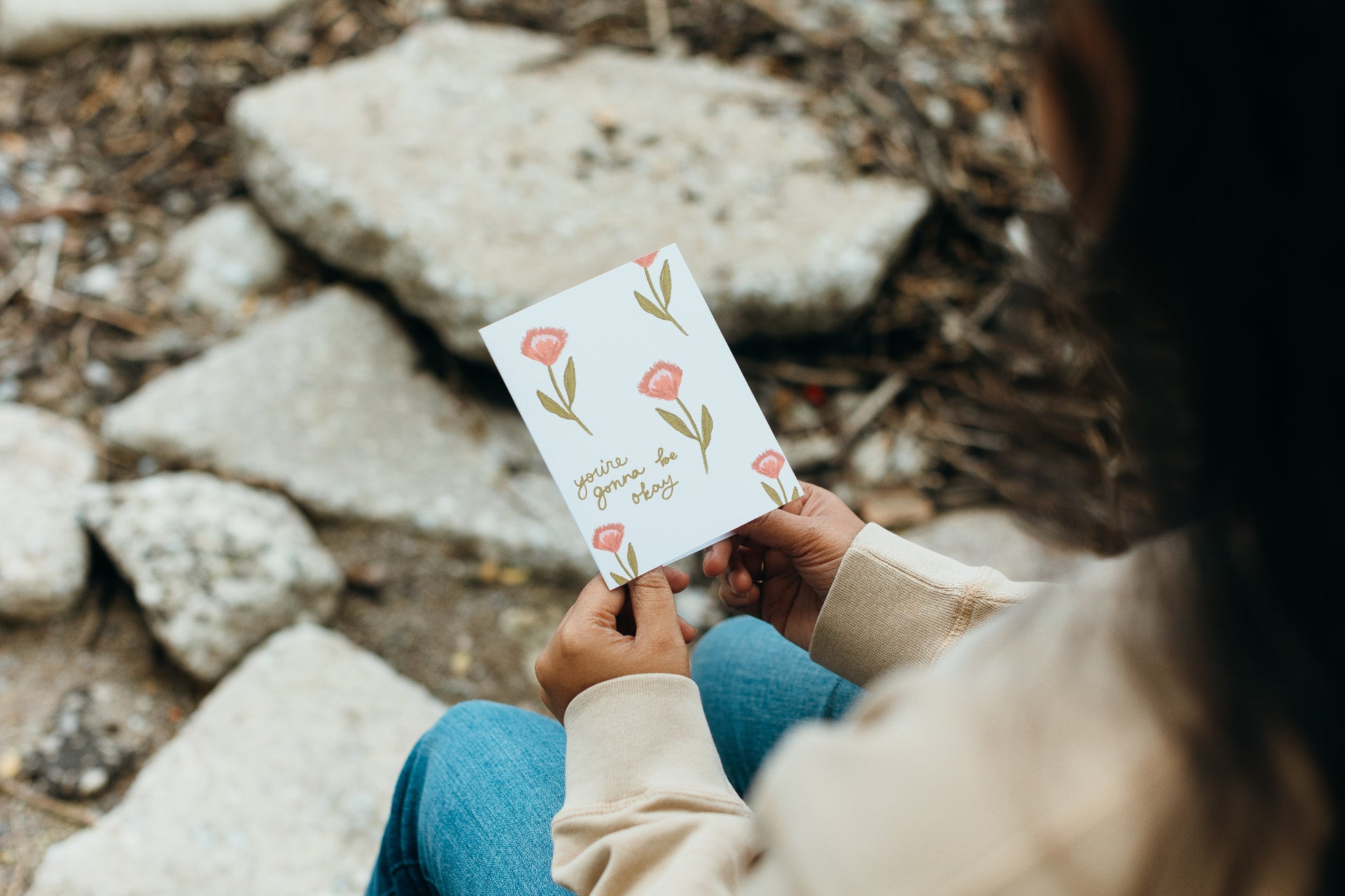 A person sitting outdoors on stone holds a small white card with pink flower illustrations and the phrase 'you are enough' in script.