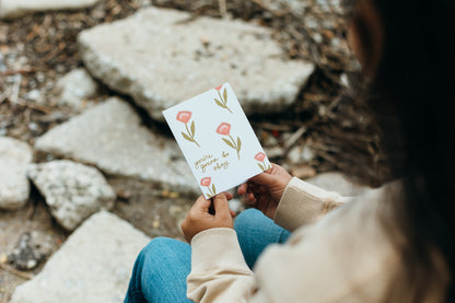 A person sitting outdoors on stone holds a small white card with pink flower illustrations and the phrase 'you are enough' in script.