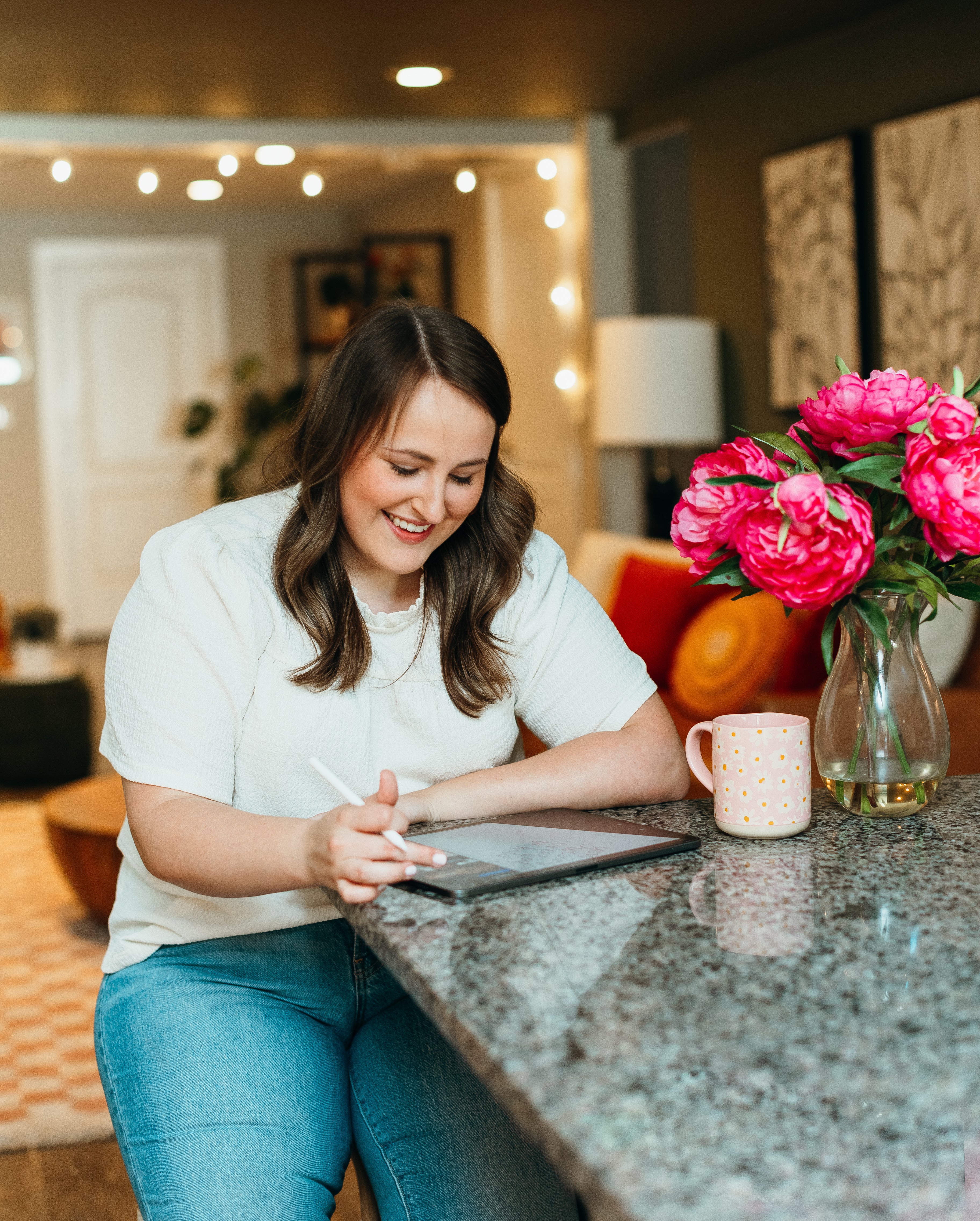 Woman drawing and using a tablet at a counter with pink flowers and a mug in the background